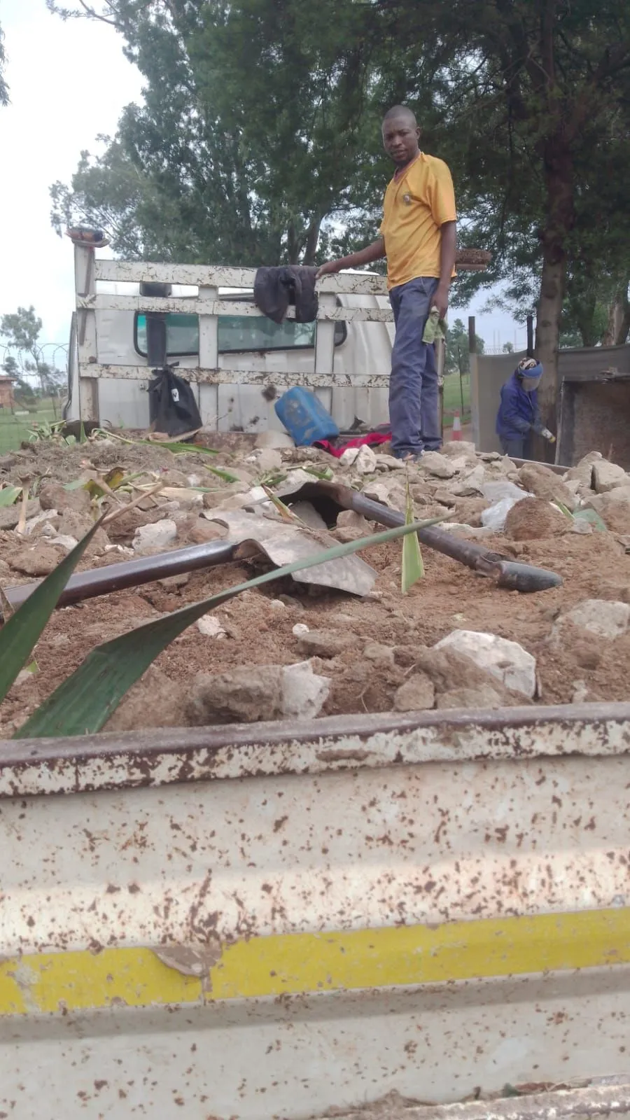 Crew member standing on a truck load of soil, rocks and pipe offcuts, with a second worker behind